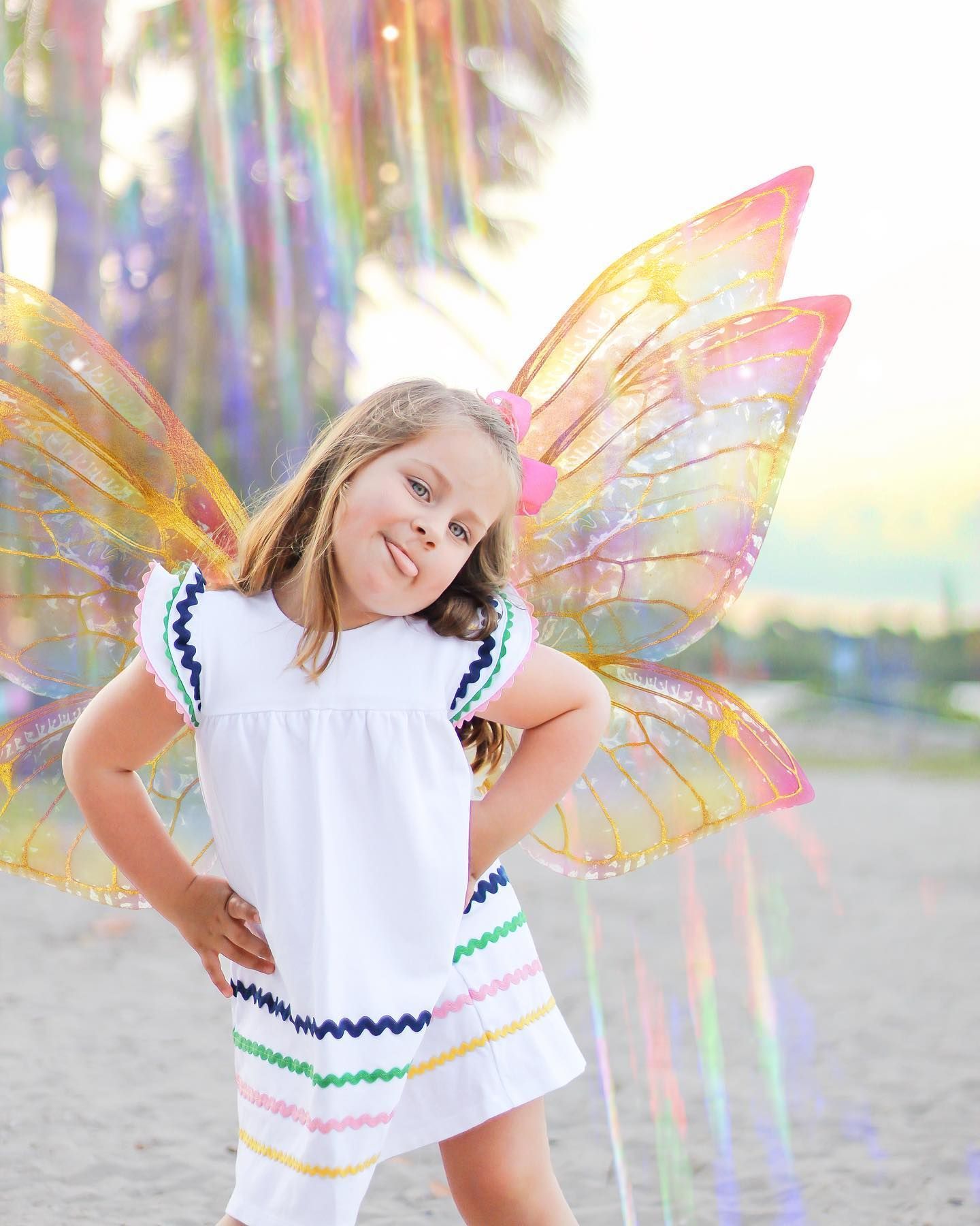 A child in a white dress with decorative butterfly wings stands playfully amid iridescent lighting and palm tree at Dubois inlet beach.