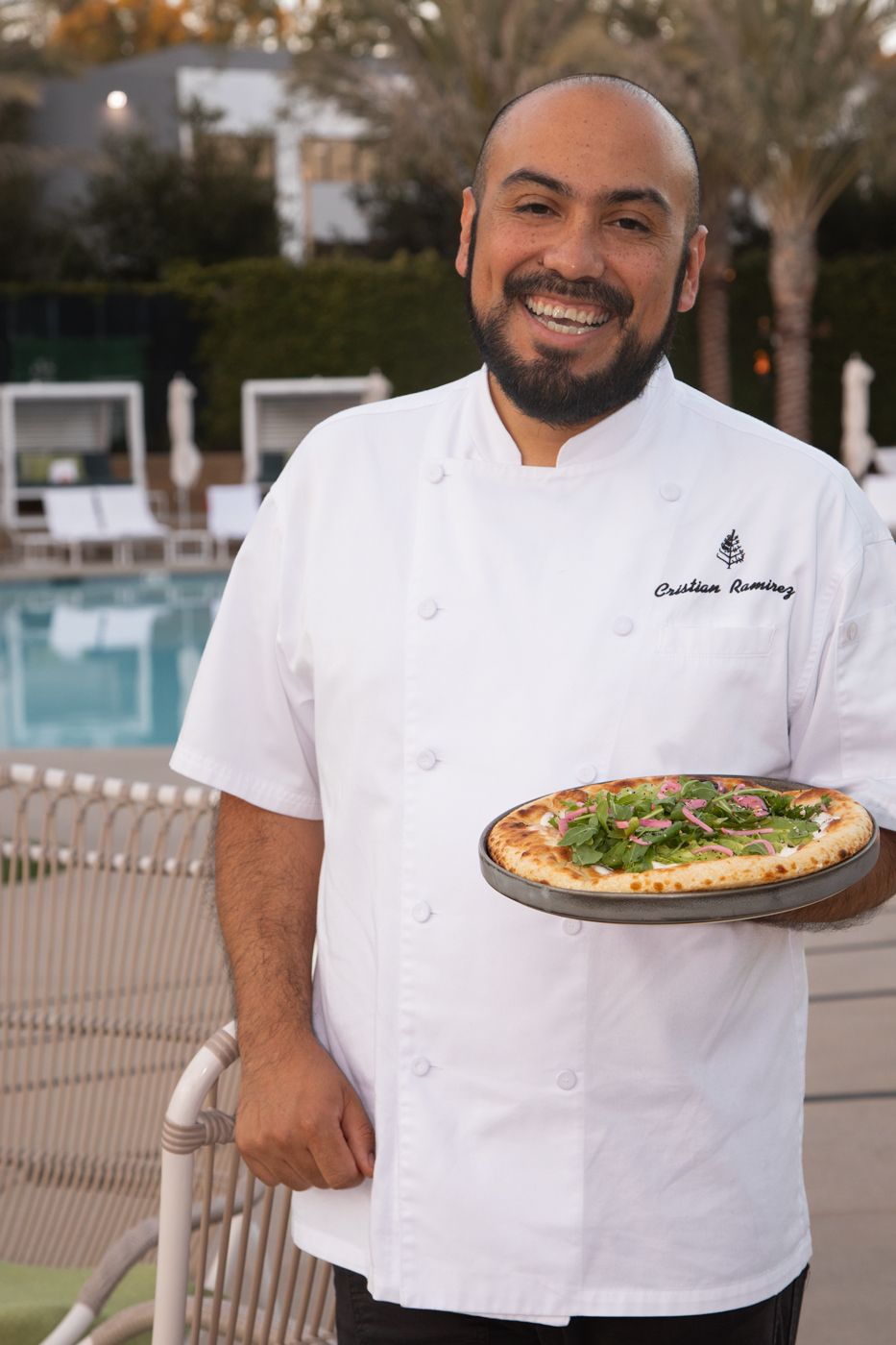 Chef in a chegs coat holding a dish at a hotel patio and smiling