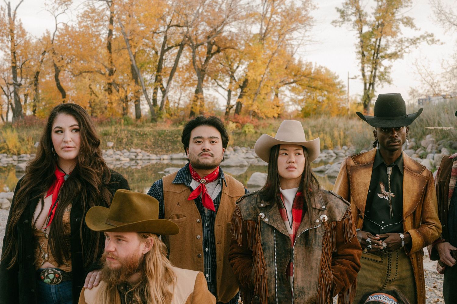 A group wearing western-style clothing with cowboy hats and leather jackets stands together against autumn trees.