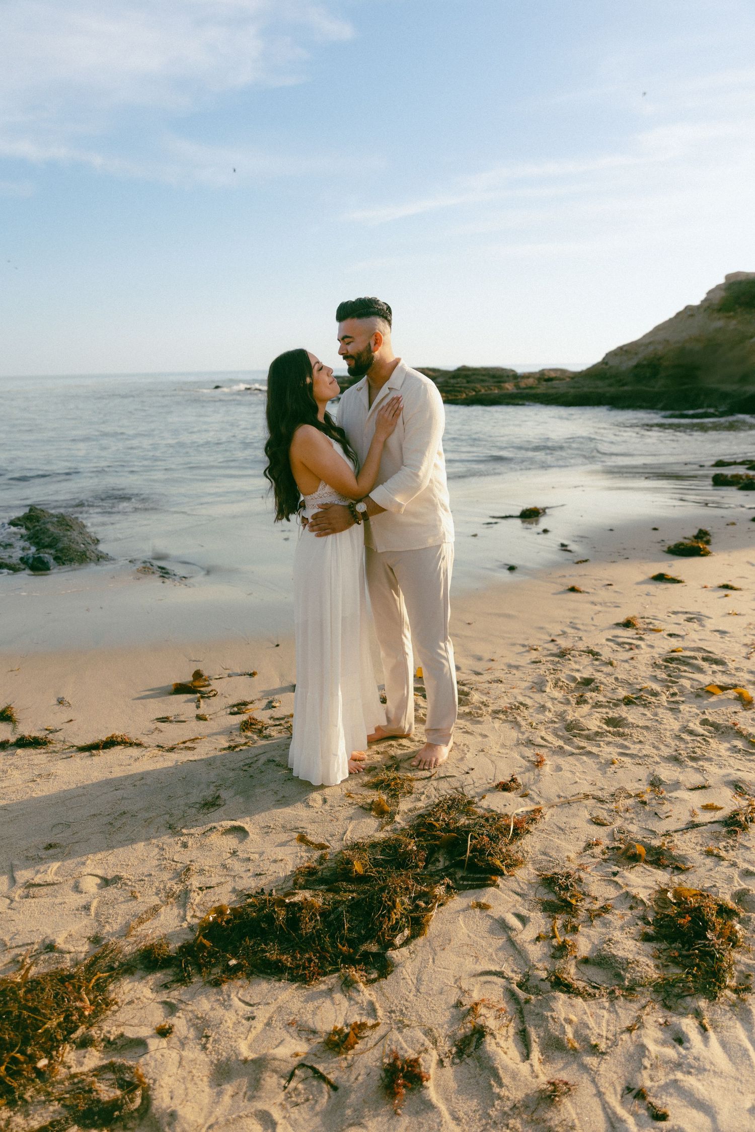 A romantic couple embraces on a scenic beach with rocky cliffs and blue ocean waters in the background.