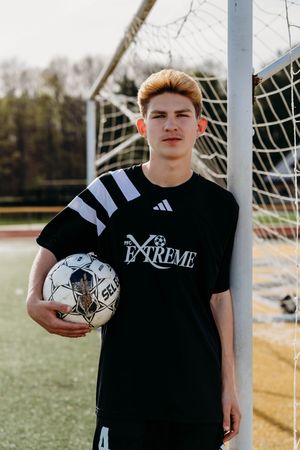 A young athlete in a black t-shirt poses by a soccer goal with a soccer ball on a sunny day at a sports field.