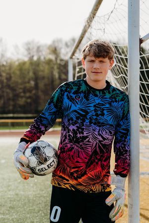 Soccer goalkeeper in colorful tropical print jersey stands by goal post holding ball on turf field.