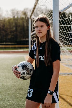 Soccer player in black uniform holding ball stands in front of goal net on turf field during photo session.
