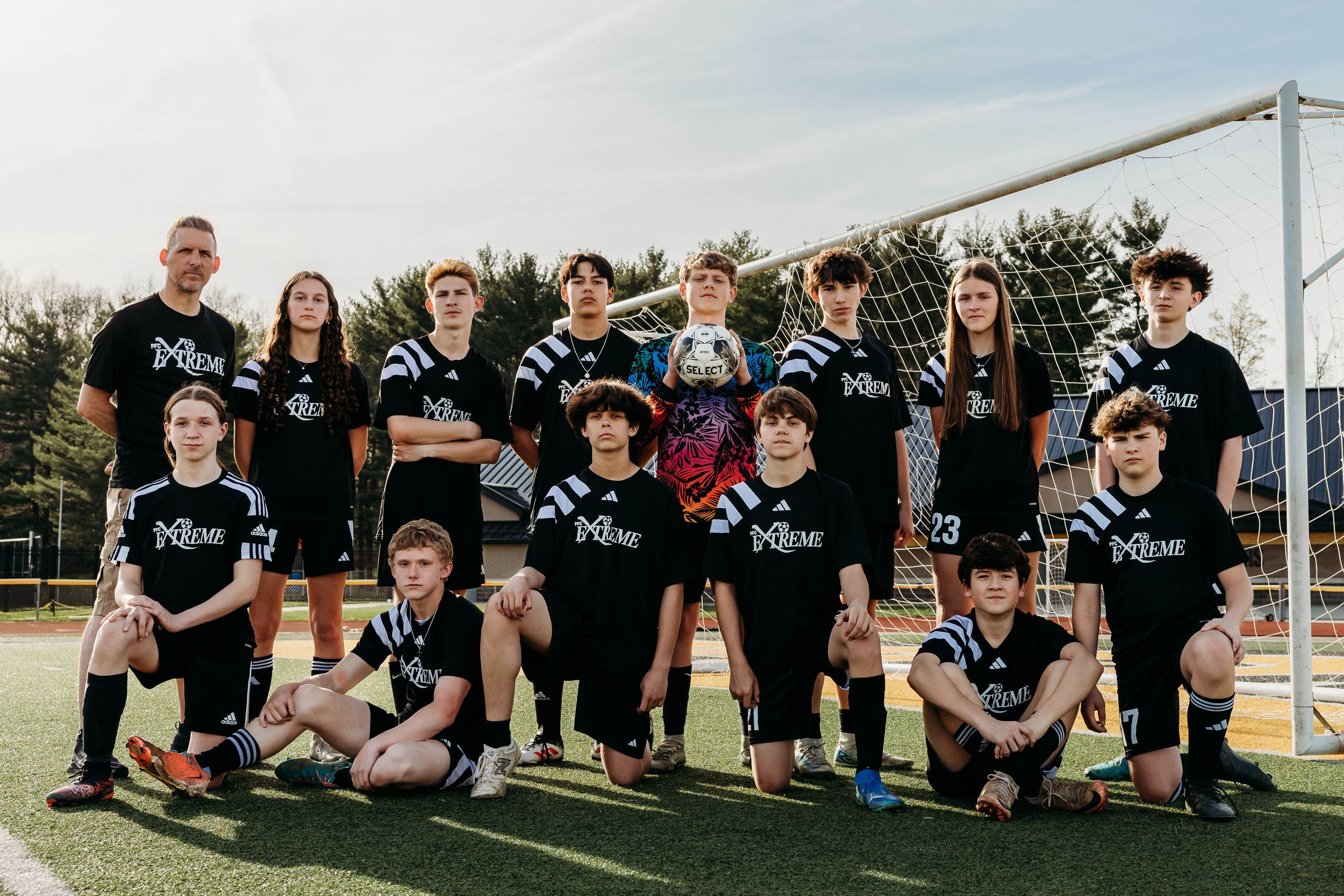 A youth soccer team poses together on a field wearing matching black uniforms in front of a goal post at sunset.