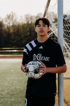 A young soccer player in a black uniform holds a Select brand soccer ball next to a goal post on a grass field.