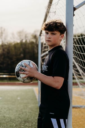 Young soccer player in black jersey holds a ball while leaning against goal post on sports field at sunset.