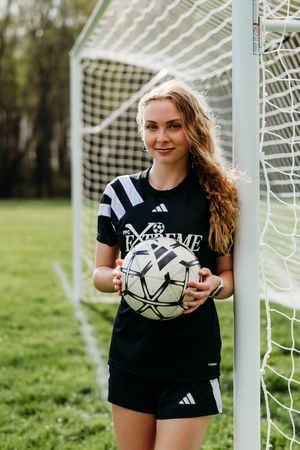 Soccer player in Adidas black uniform holding a ball while standing next to goal post on grass field.