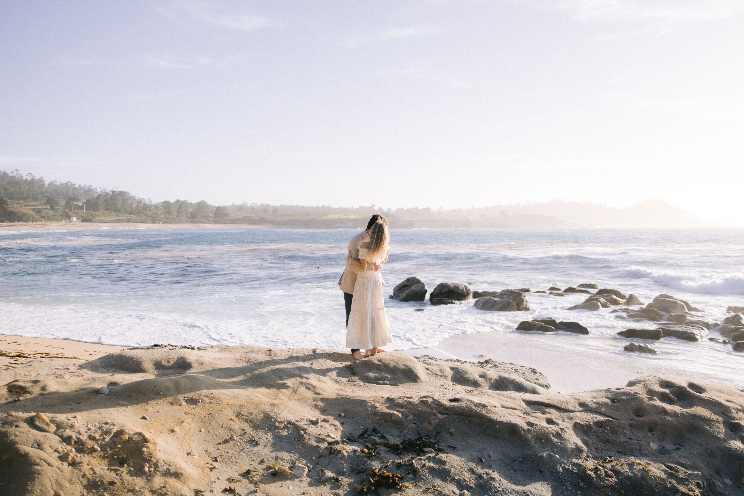 A white-clad figure stands on rocky oceanfront cliffs during golden hour as gentle waves lap against the shore.