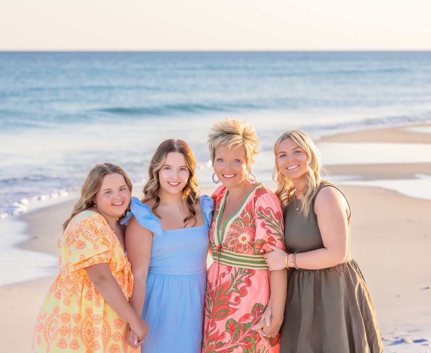Four friends in colorful sundresses pose together smiling on a sunny beach with ocean waves in the background.