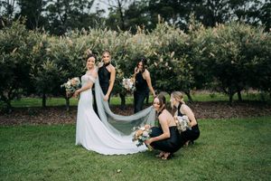 Bridesmaids in black dresses pose with floral bouquets on a lush green lawn.