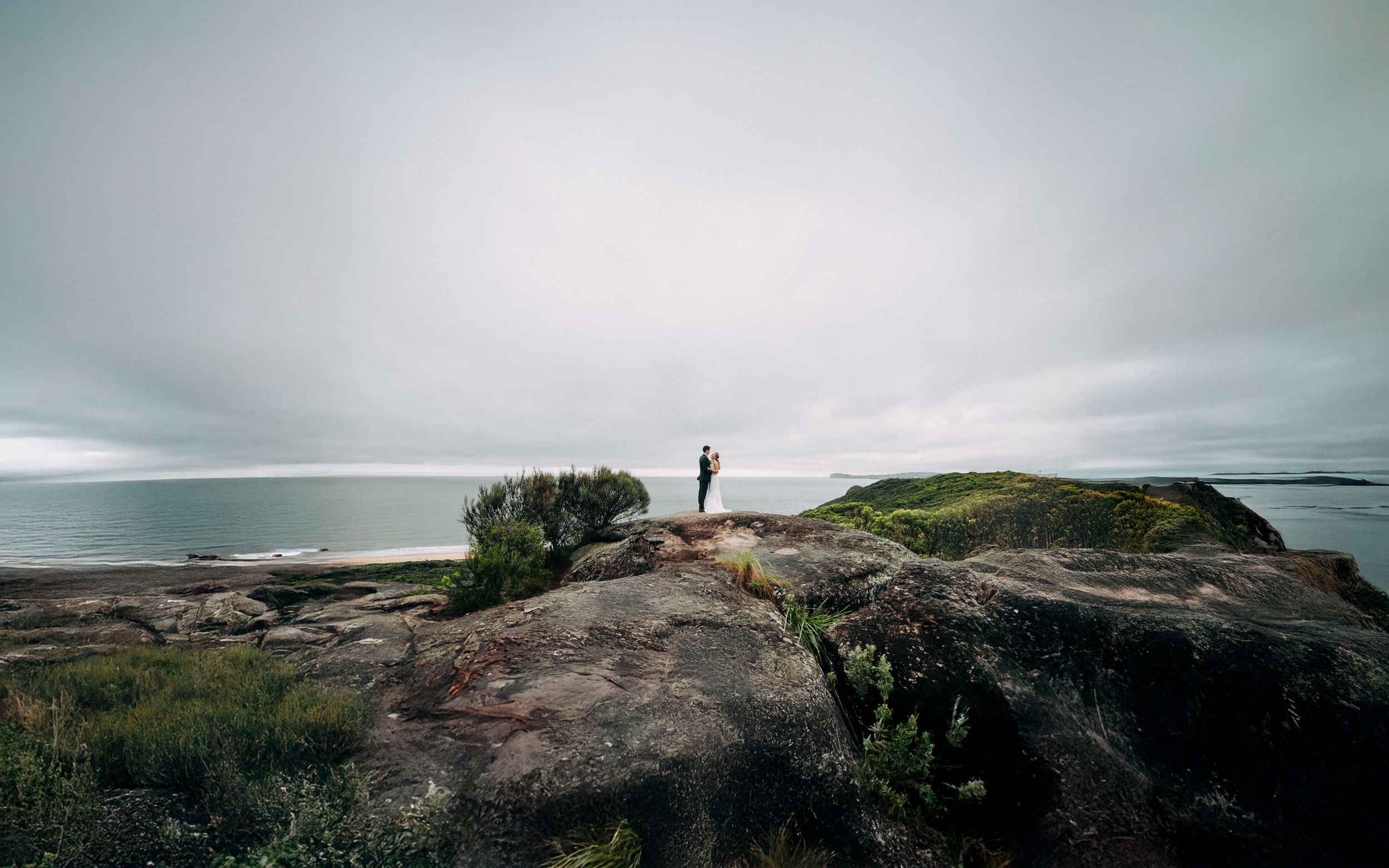 Two figures stand on dramatic rocky coastal cliffs overlooking the ocean.
