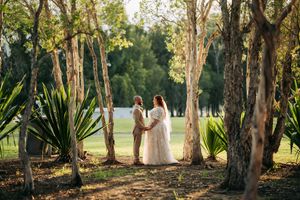 A romantic wedding moment captured between trees and tropical plants in a sunlit garden setting.