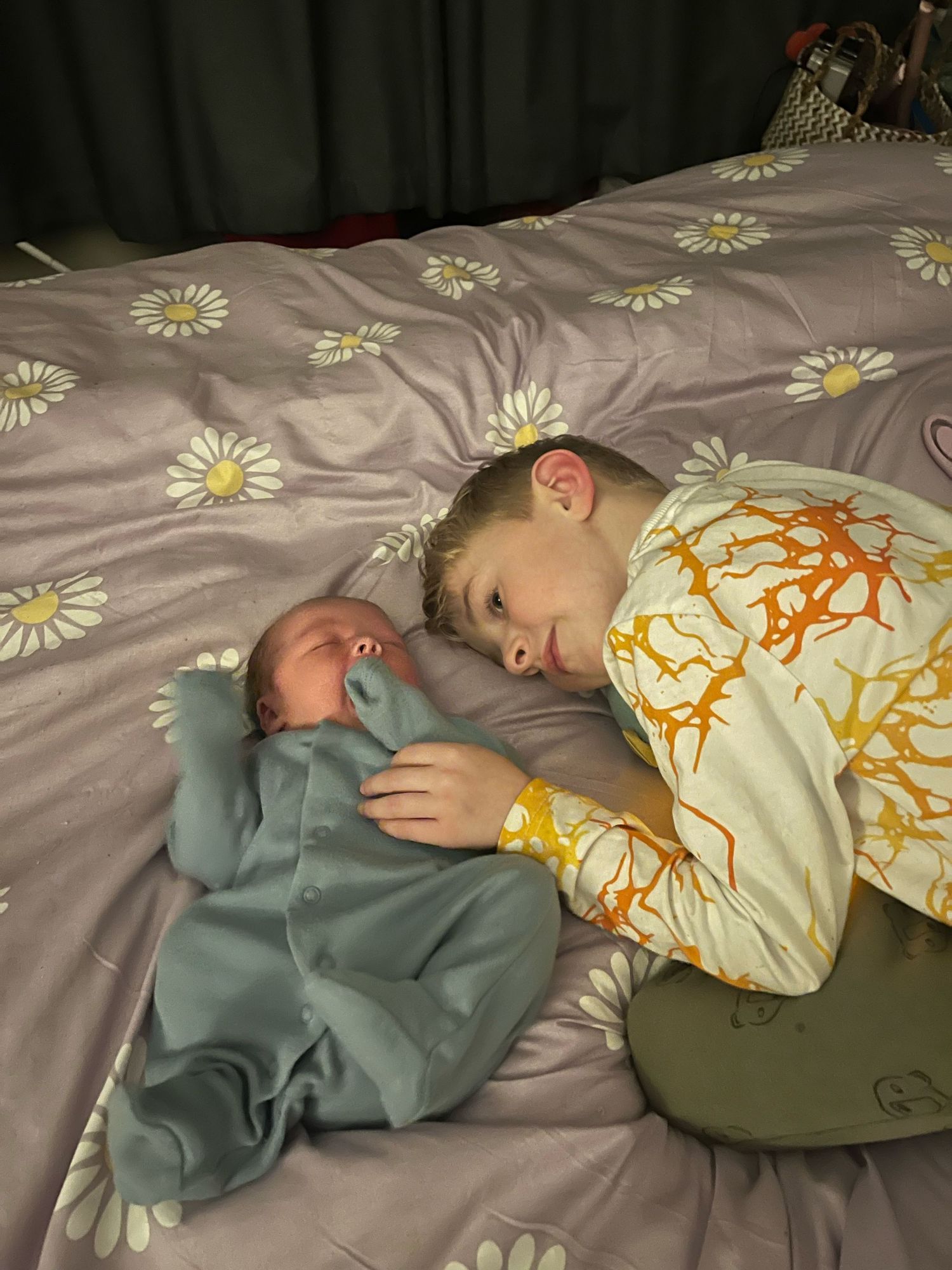 Two people sleeping peacefully on a gray floral patterned bedspread, one in a mint green outfit and one in yellow.