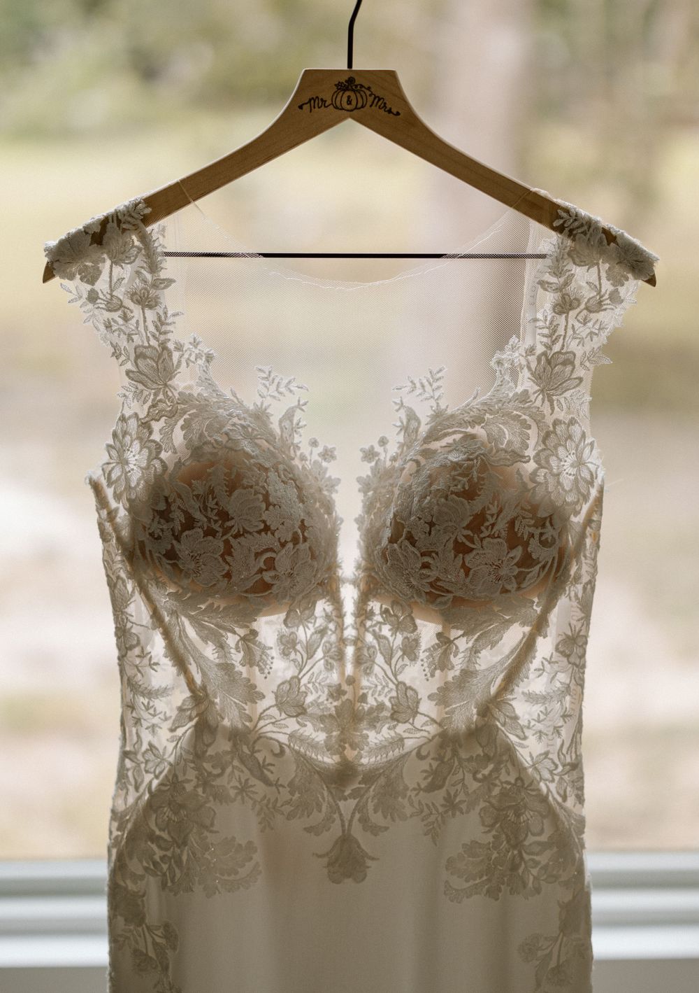 White lace wedding dress bodice with intricate floral details hanging on wooden hanger against window light.