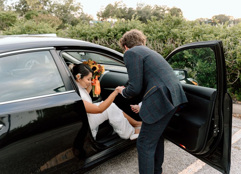 A person assists another person getting into a black car parked on a road with greenery in the background.