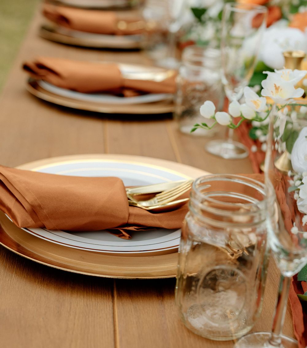 Elegant table setting with peach napkins, white plates, mason jars and delicate floral centerpieces on a wooden surface.
