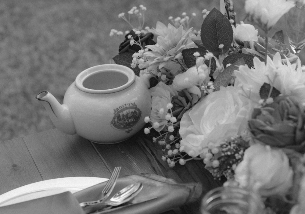 A white ceramic teapot sits beside a floral arrangement with white blooms on a wooden table in black and white.