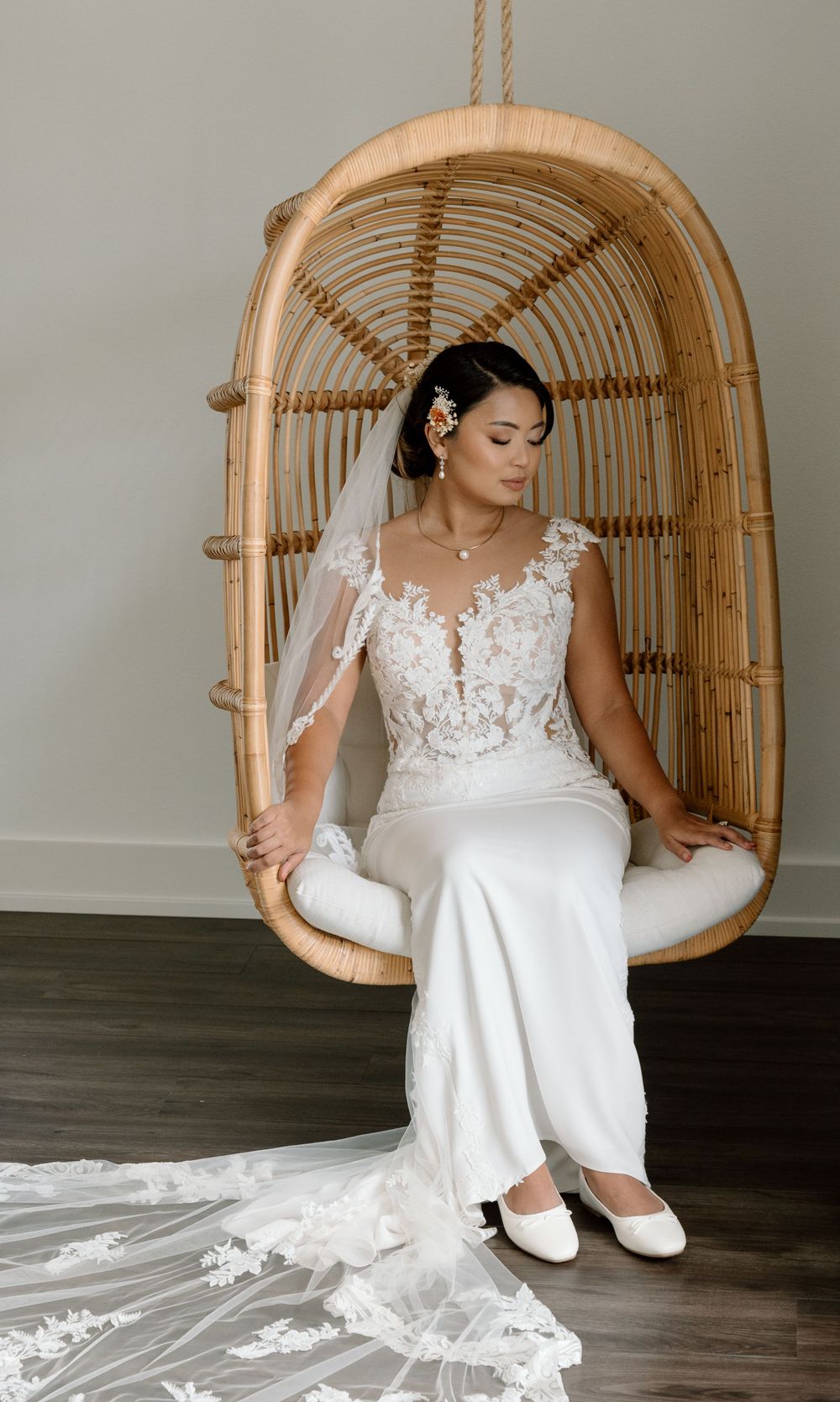 Elegant bride in lace wedding dress and white flats sitting in rattan egg chair with flowing train.