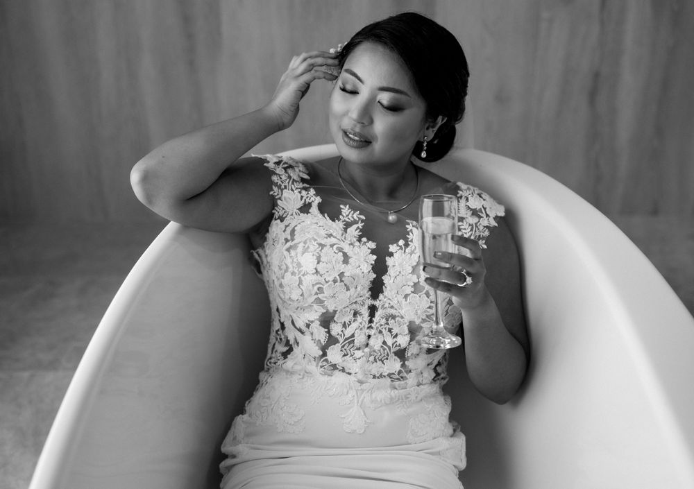 A person in a wedding dress relaxes in a bathtub while holding a drink glass in black and white photography.