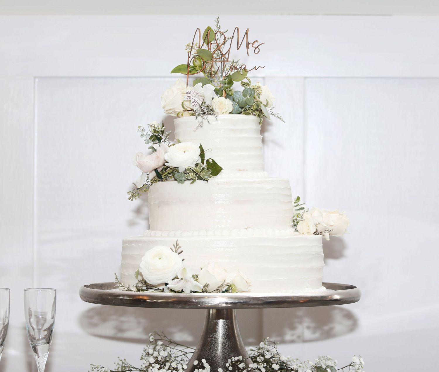 An elegant white wedding cake decorated with fresh flowers and greenery on a silver cake stand.