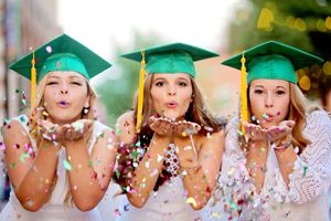 Three graduates in green caps and gowns blow glitter confetti in celebration.