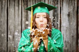 Graduate in green cap and gown blowing gold glitter confetti against rustic wooden background.