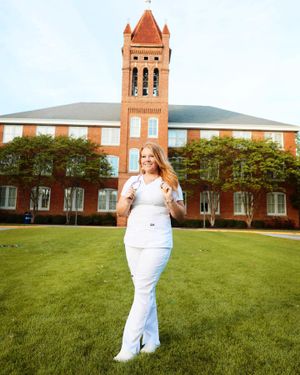 Graduate in white dress poses on lawn in front of brick university building with bell tower.