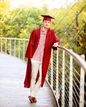 Graduate in red cap and gown walking on modern bridge during golden hour.