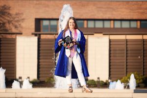 Graduate in blue cap and gown holding bouquet in front of brick building with white fountains.