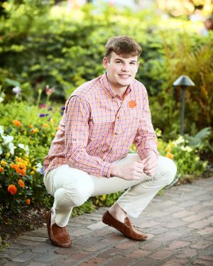 A student in a red checkered shirt and khakis crouches on a garden path surrounded by colorful flowers.