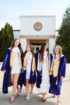 Four graduates in blue caps and gowns with gold stoles stand laughing outside Belton one path building entrance.