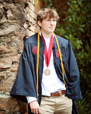 Graduate in dark academic regalia poses against stone wall wearing gold and red honor cords and medallion.