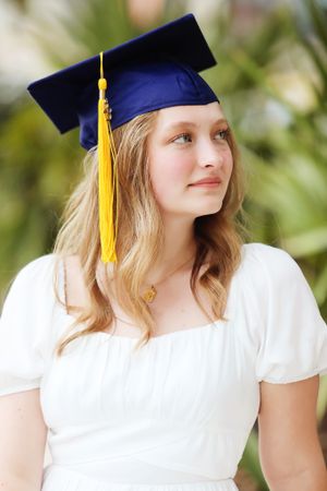 Graduate in navy cap with gold tassel and white dress poses for portrait outdoors.