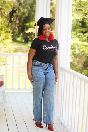 Graduate in Carolina shirt and cap poses on white porch in casual attire.