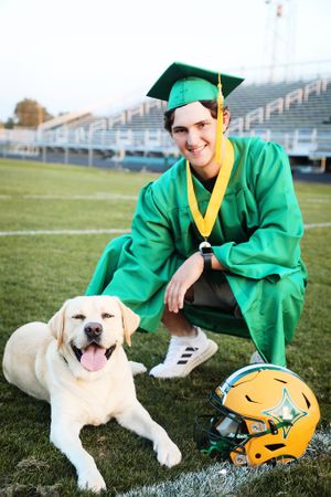 Graduate in green gown poses with happy yellow Labrador retriever next to football helmet on field.