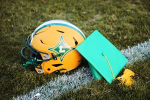 Gold football helmet and green graduation cap rest on white field marking line.