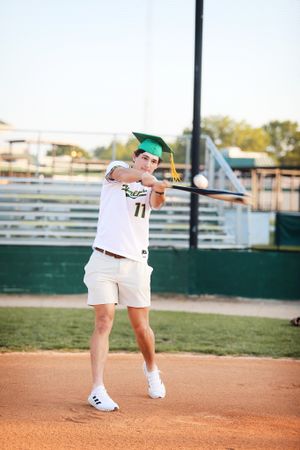 Graduate in baseball uniform poses with batting stance on baseball field.