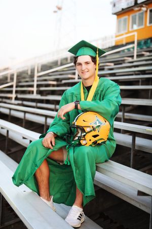 Graduate in green gown sits on stadium bleachers holding gold football helmet.