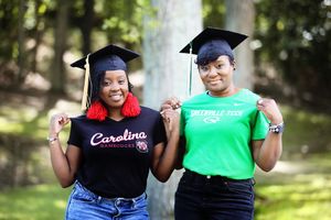 Two graduates in caps celebrate together wearing Carolina and athletic apparel.