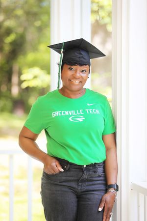 Graduate in Greenville Tech shirt poses by window in graduation cap.