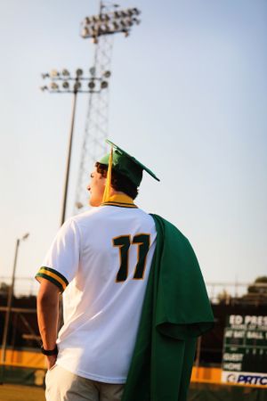 Baseball player in green graduation cap wearing jersey number 11 stands beneath stadium lights at dusk.