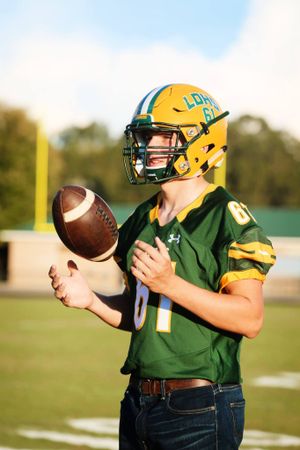 Football player in green and yellow uniform poses with ball on field at sunset.
