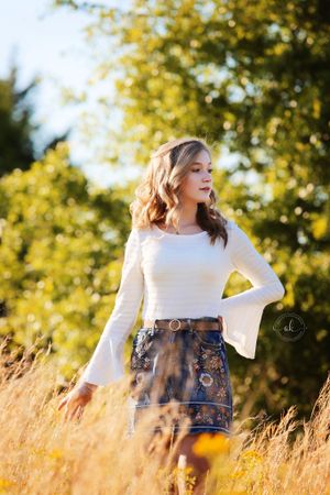 Portrait of person in white bell-sleeve top and patterned skirt standing in golden field at sunset.