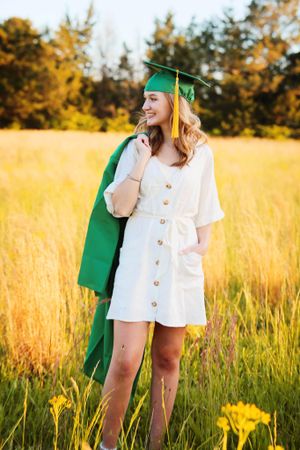 Graduate in white dress and green cap and gown poses in golden wheat field at sunset.