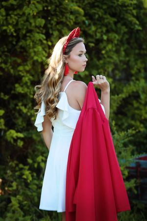 A white summer dress with red accessories styled against lush greenery in an outdoor portrait setting.
