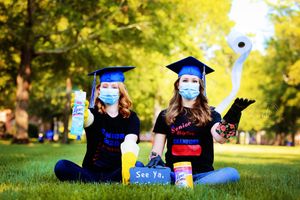 Two graduates in blue caps and gowns wearing face masks celebrate outdoors with diplomas.