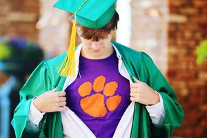 A person in a green graduation gown reveals a purple shirt with an orange paw print logo underneath.