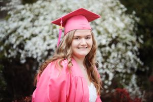 Graduate in pink cap and gown poses with flowering trees in background.