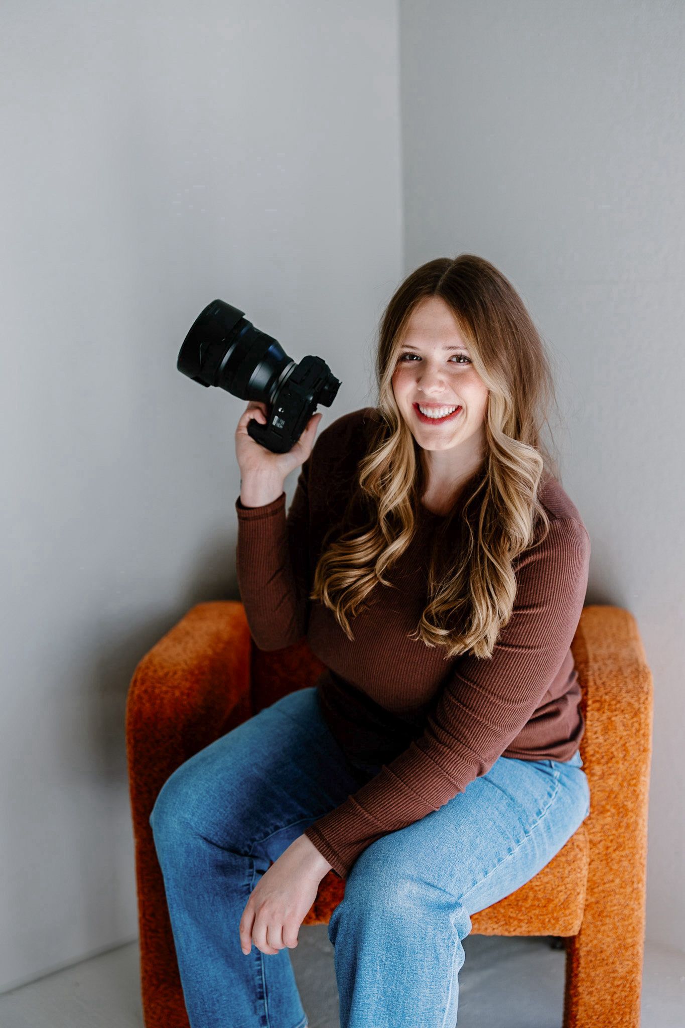 Professional photographer sitting in an orange chair holding a DSLR camera against a gray wall.