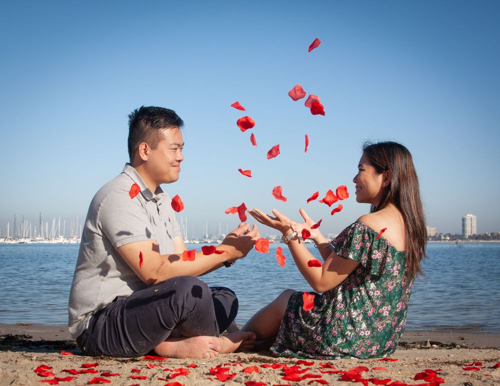 Couple sitting by waterfront throwing red rose petals in the air during romantic moment.